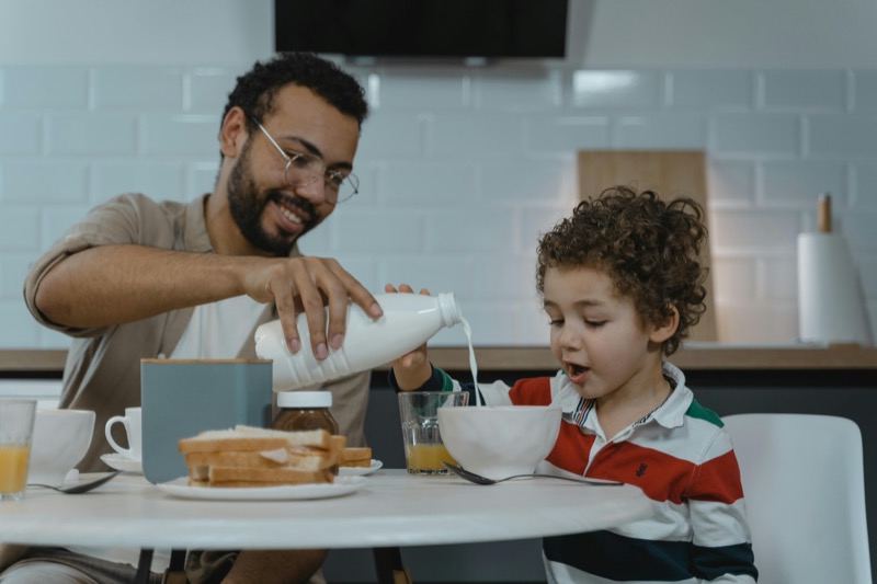 Father and son sharing breakfast together at kitchen table, father pouring milk into boy's bowl